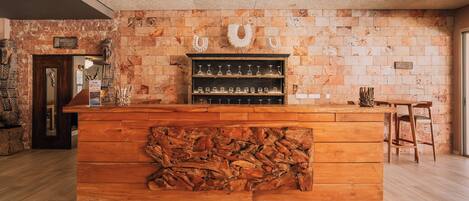 A wooden bar with a rustic design, a stone wall, and a shelf with glasses.