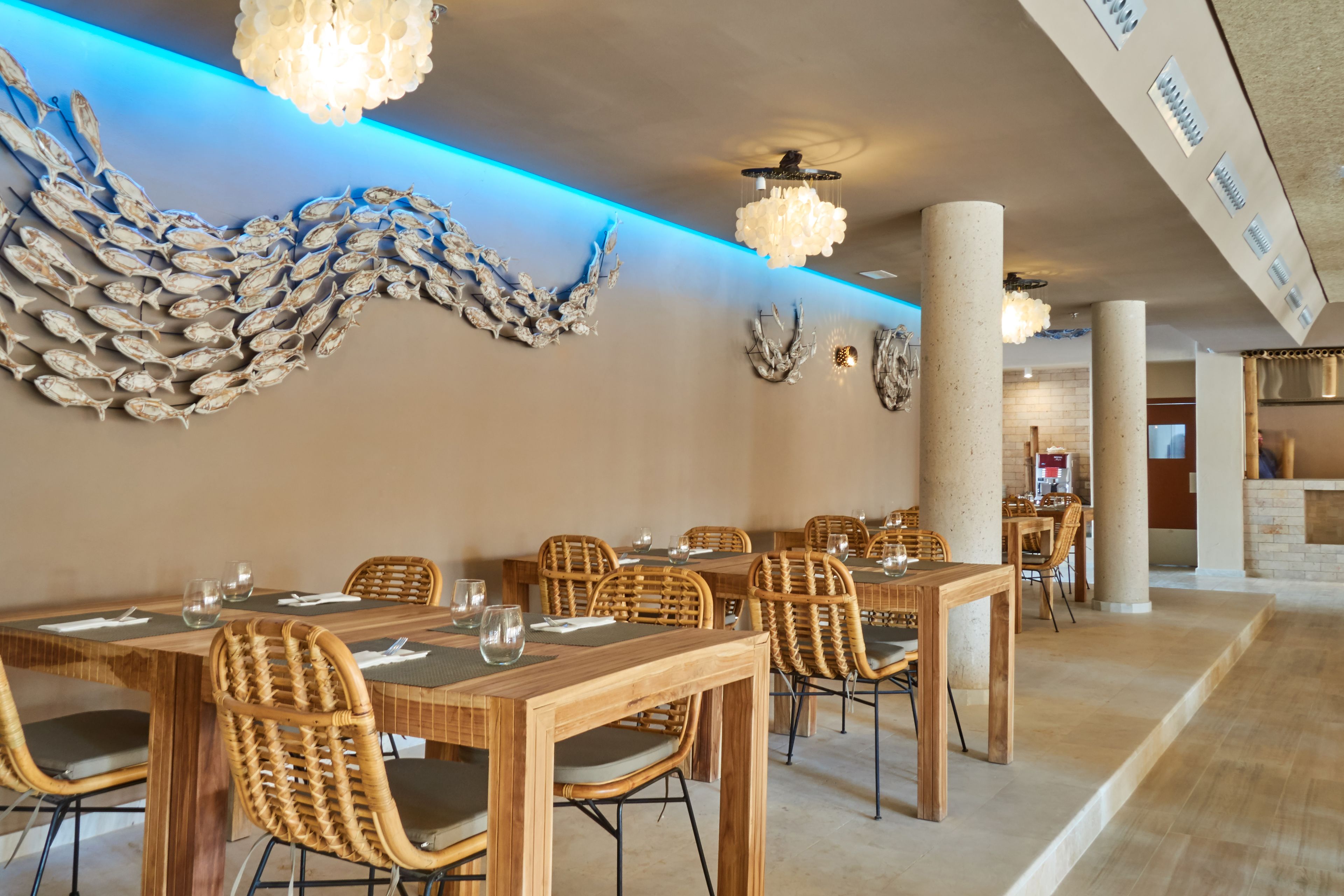 A dining area with wooden tables and wicker chairs, a decorative fish wall art, and a blue-lit ceiling.