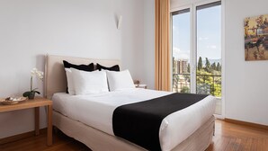 Un dormitorio moderno con una cama grande, una mesita de noche de madera, una orquídea en maceta y vistas a un paisaje urbano a través de un ventanal amplio.