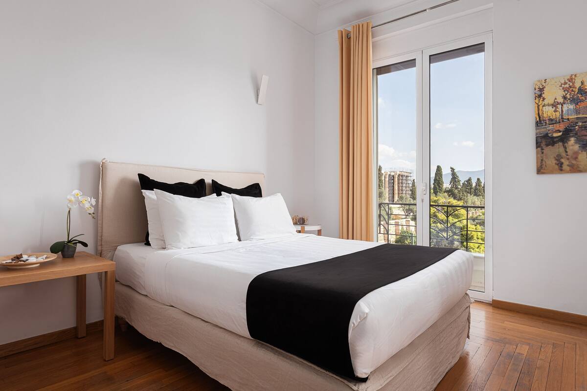 A modern bedroom with a large bed, a wooden nightstand, a potted orchid, and a view of a cityscape through a large window.