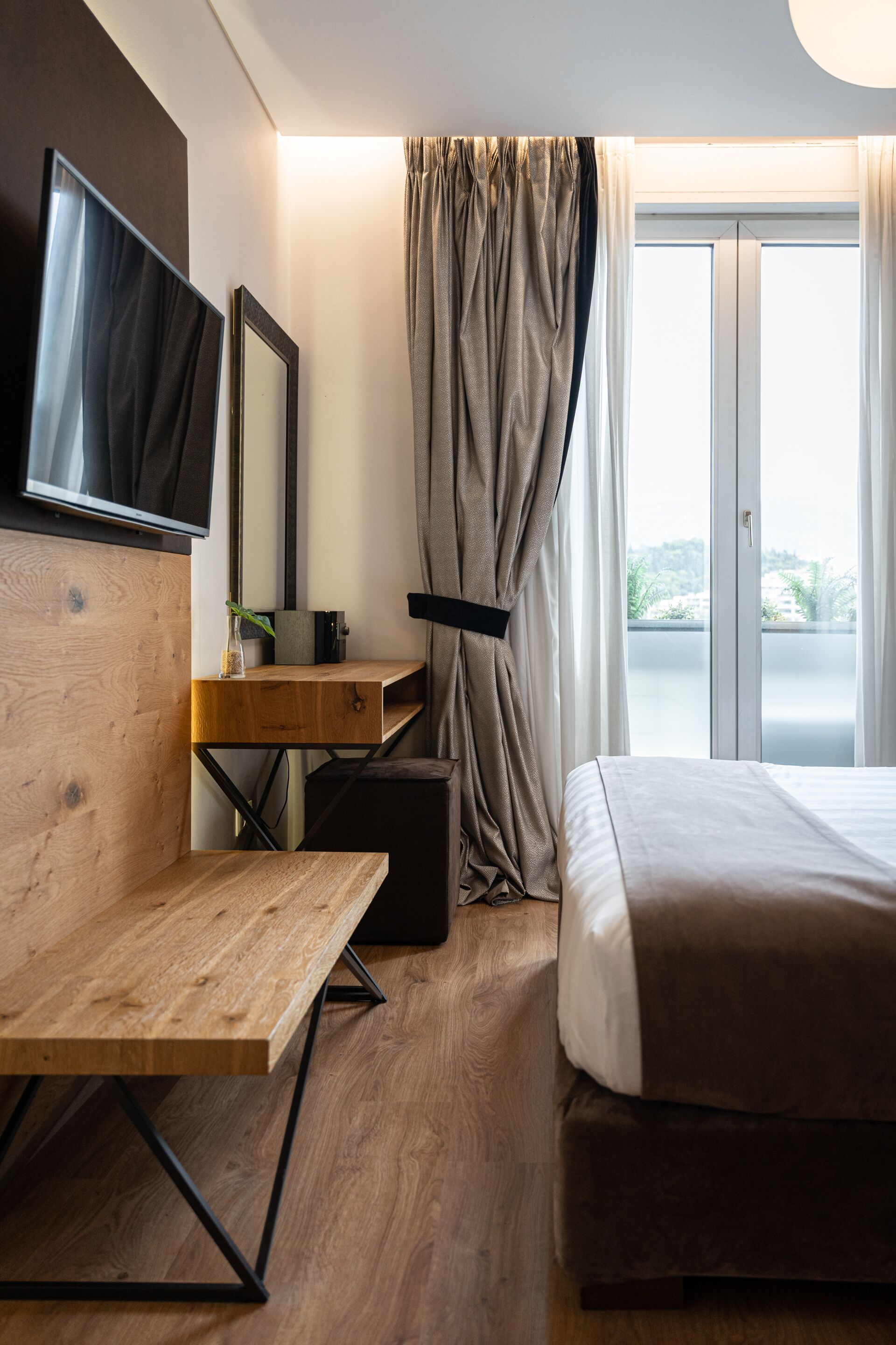 Ein Hotelzimmer mit Schreibtisch aus Holz, einem Bett, einem an der Wand montierten Fernseher und Blick ins Freie durch ein großes Fenster.