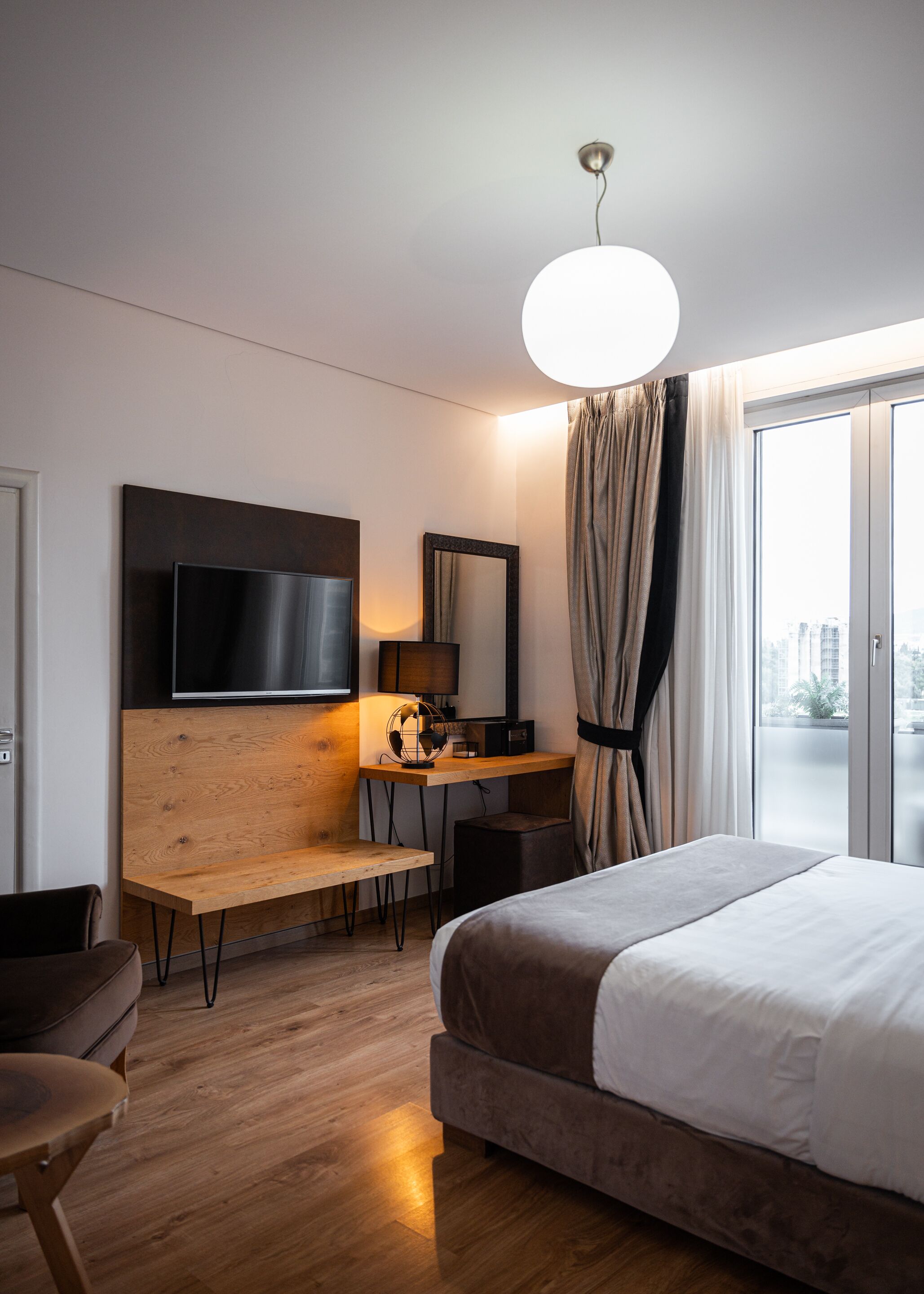 A modern hotel room with a large bed, a desk with a computer, a flat-screen TV, and a large window with curtains.