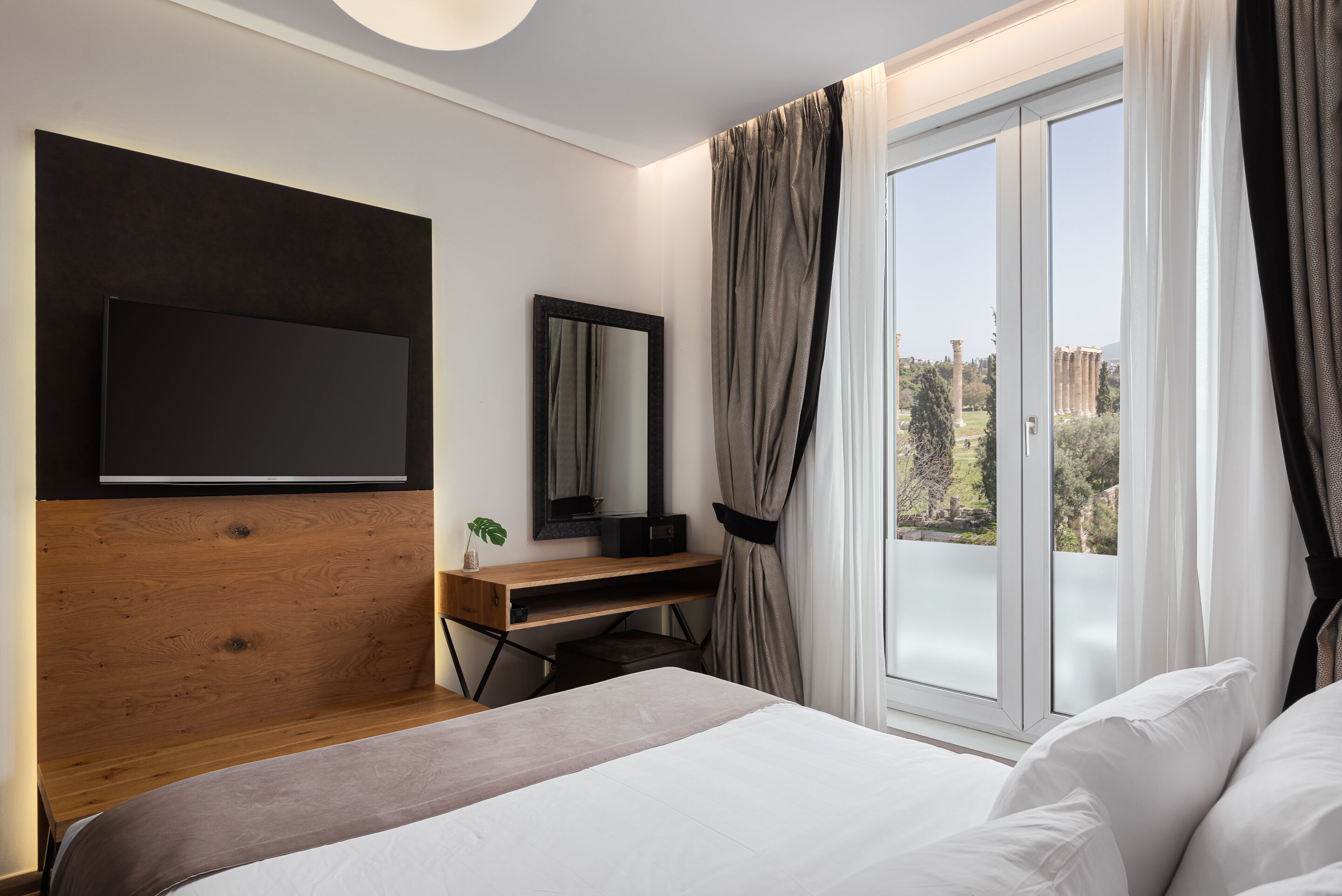 Une chambre d’hôtel moderne dotée d’un grand lit, d’un bureau en bois, d’un téléviseur à écran plat, d’un miroir et d’une grande fenêtre avec des rideaux.