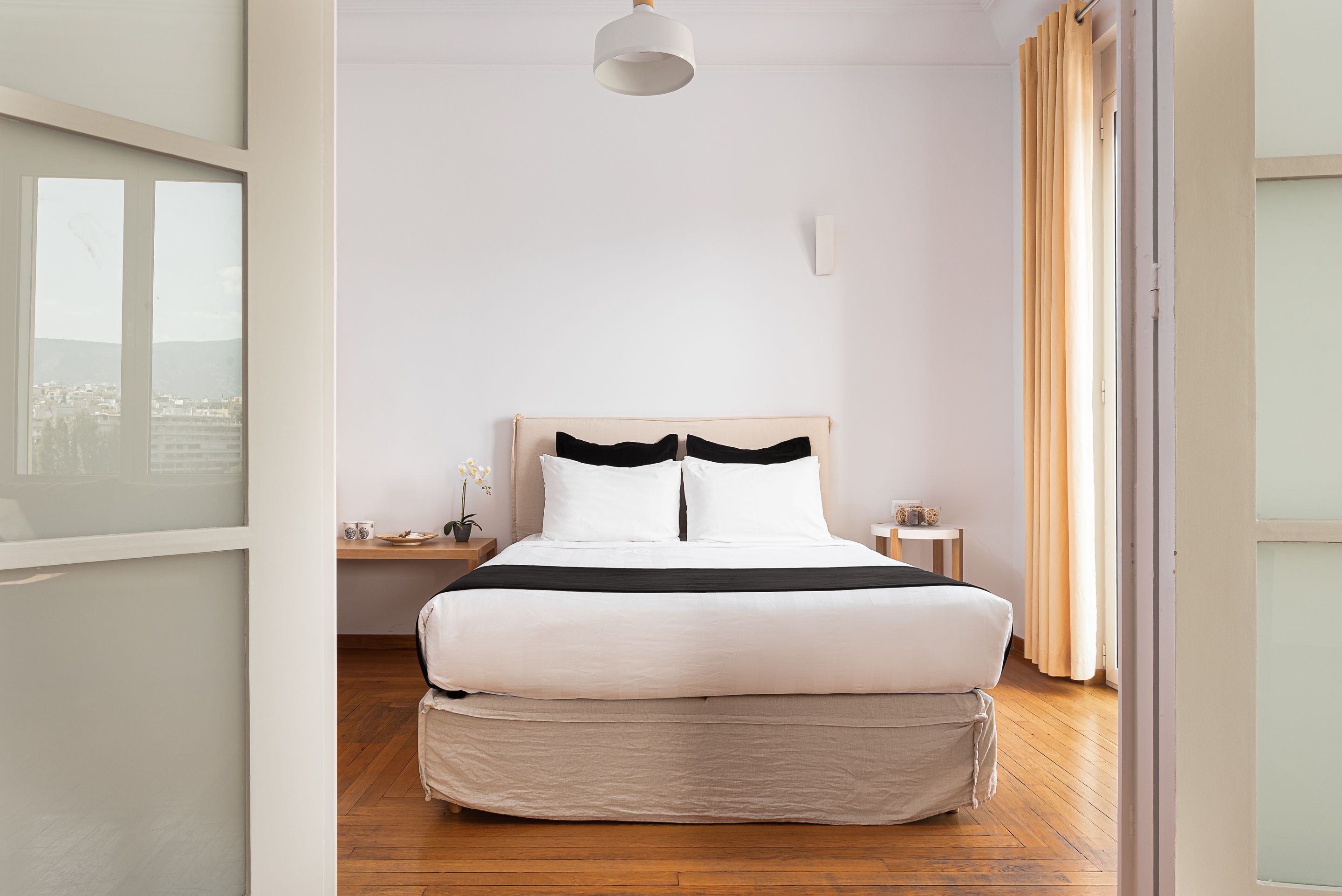 A neatly arranged bedroom with a large bed, a bedside table, a nightstand, and a view through a window.
