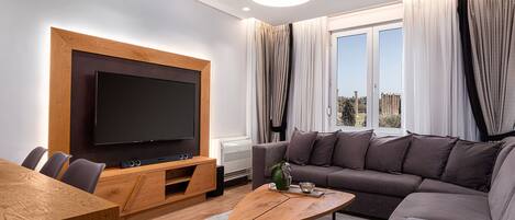 A modern living room with a flat-screen TV, wooden furniture, a gray sofa, and a large window with curtains.