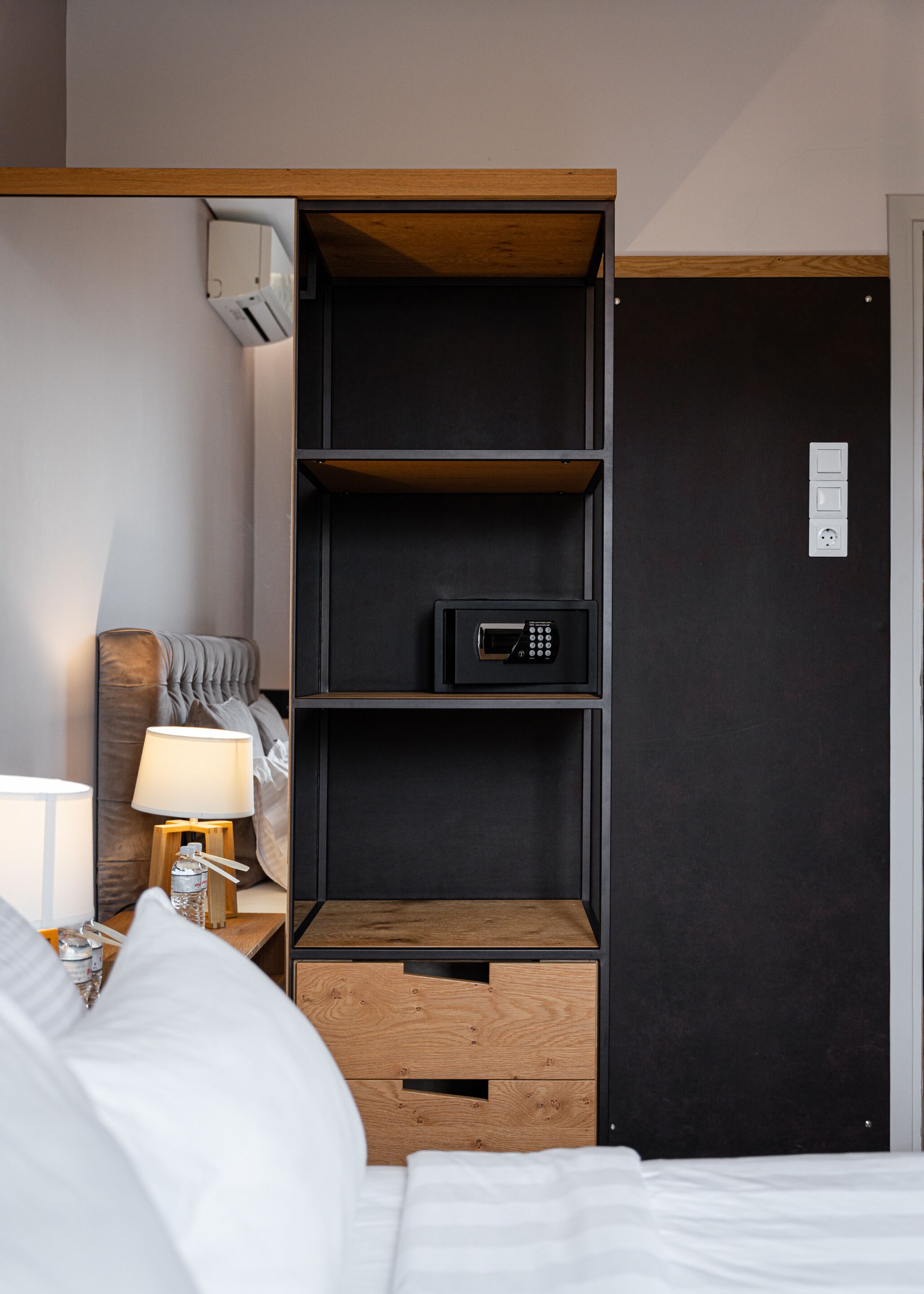 A hotel room with a bed, a nightstand with a lamp, a wooden cabinet, and a black door with a power outlet.