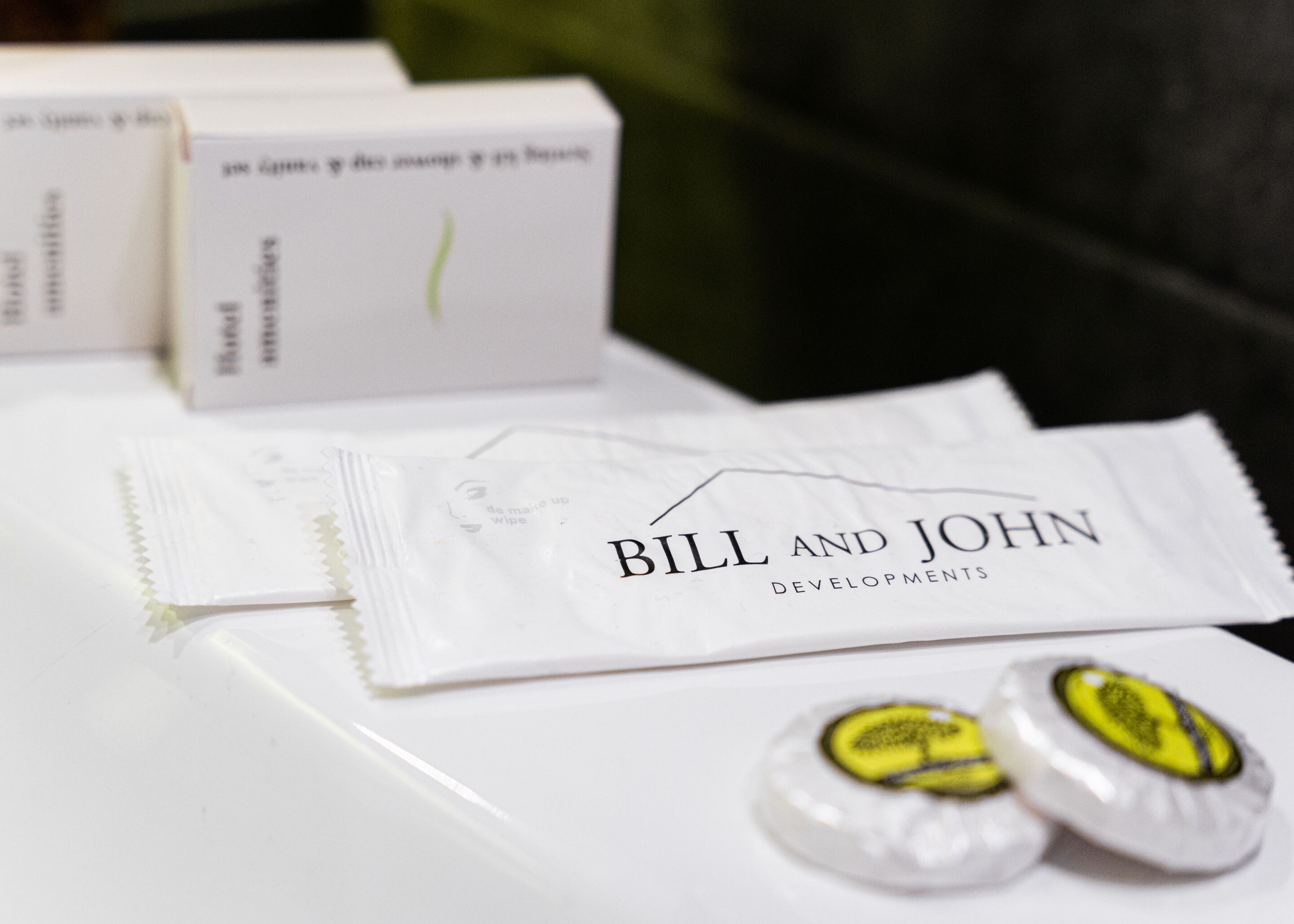 A white table with a tissue box labeled "Bill and John Developments" and two wrapped chocolates.