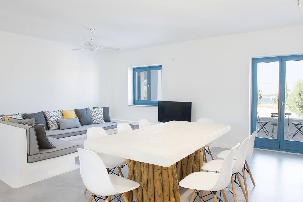 Una sala de estar moderna con una mesa blanca grande, sillas blancas, un sofá gris con cojines, un ventilador de techo y un televisor sobre un mueble.