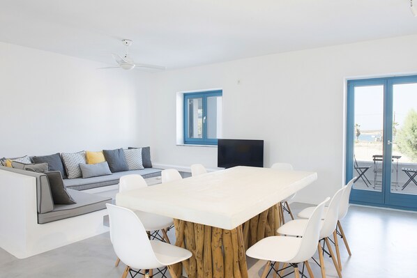 Una sala de estar moderna con una mesa blanca grande, sillas blancas, un sofá gris con cojines, un ventilador de techo y un televisor sobre un mueble.
