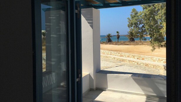 A view from a room with a glass door leading to a balcony overlooking a beach with palm trees and the sea.