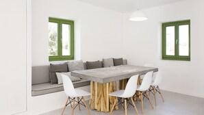 A dining area with a wooden table surrounded by white chairs, a grey sofa with cushions, and green-framed windows.