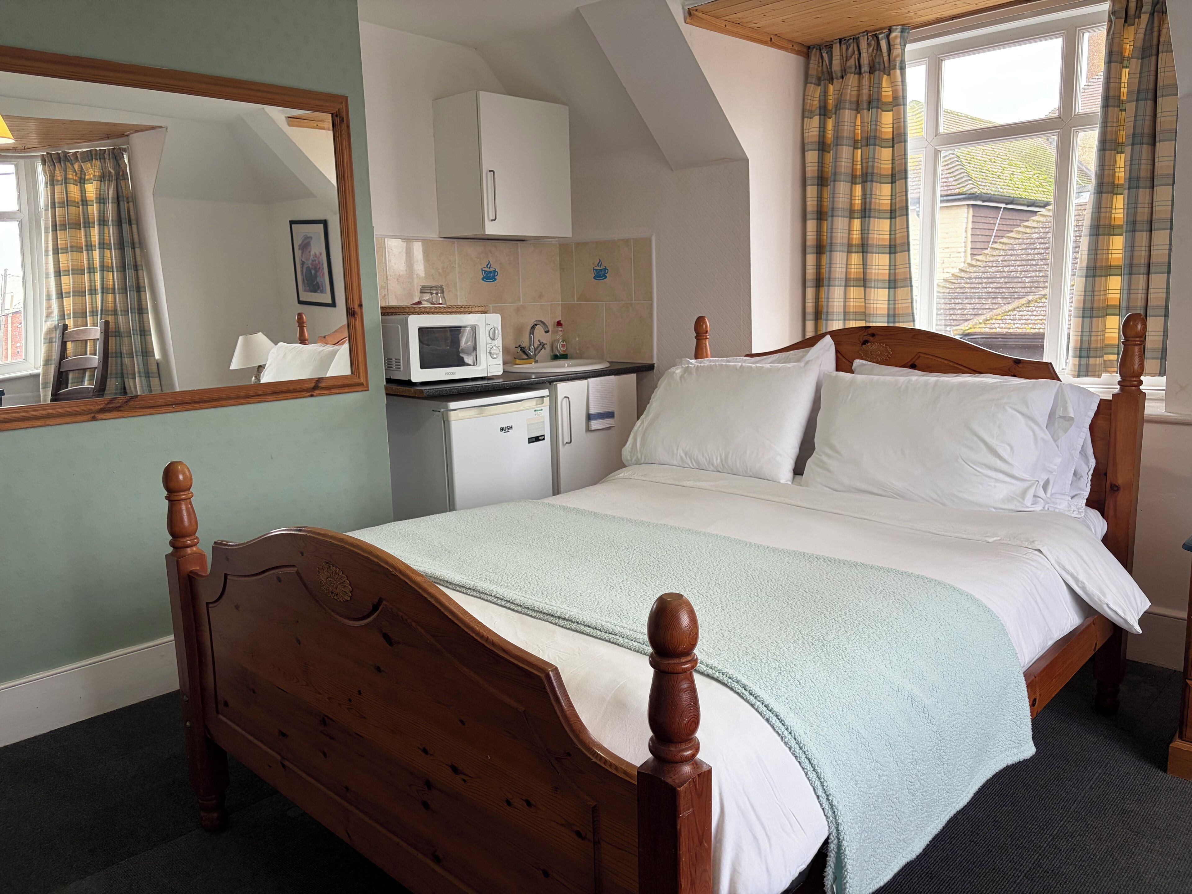 A bedroom with a wooden bed, a small kitchenette with a microwave, and a window with plaid curtains.