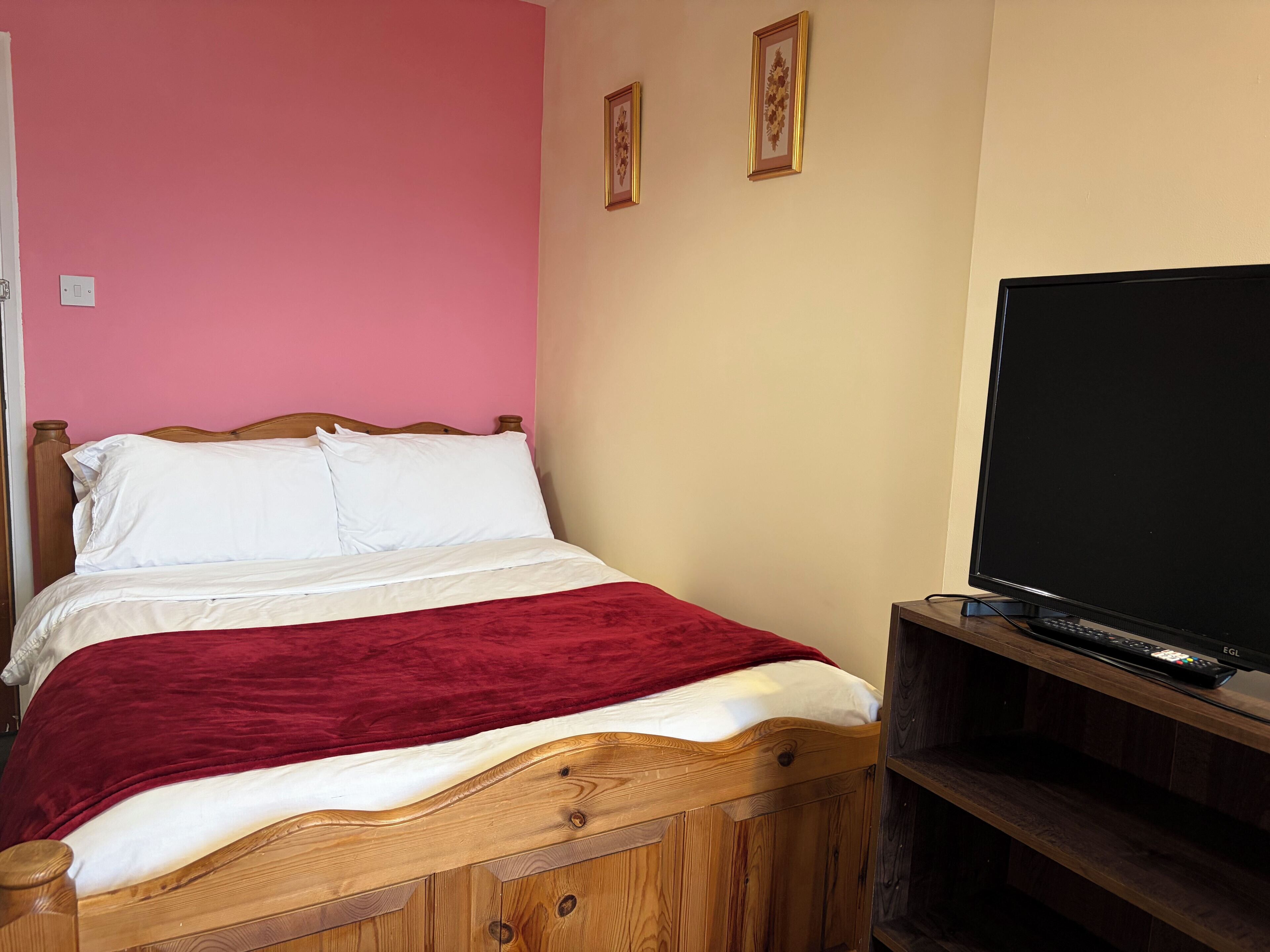 Un letto con biancheria bianca e una coperta rossa, una testiera in legno, una televisione su un mobile in legno e una parete rosa con due quadri incorniciati.