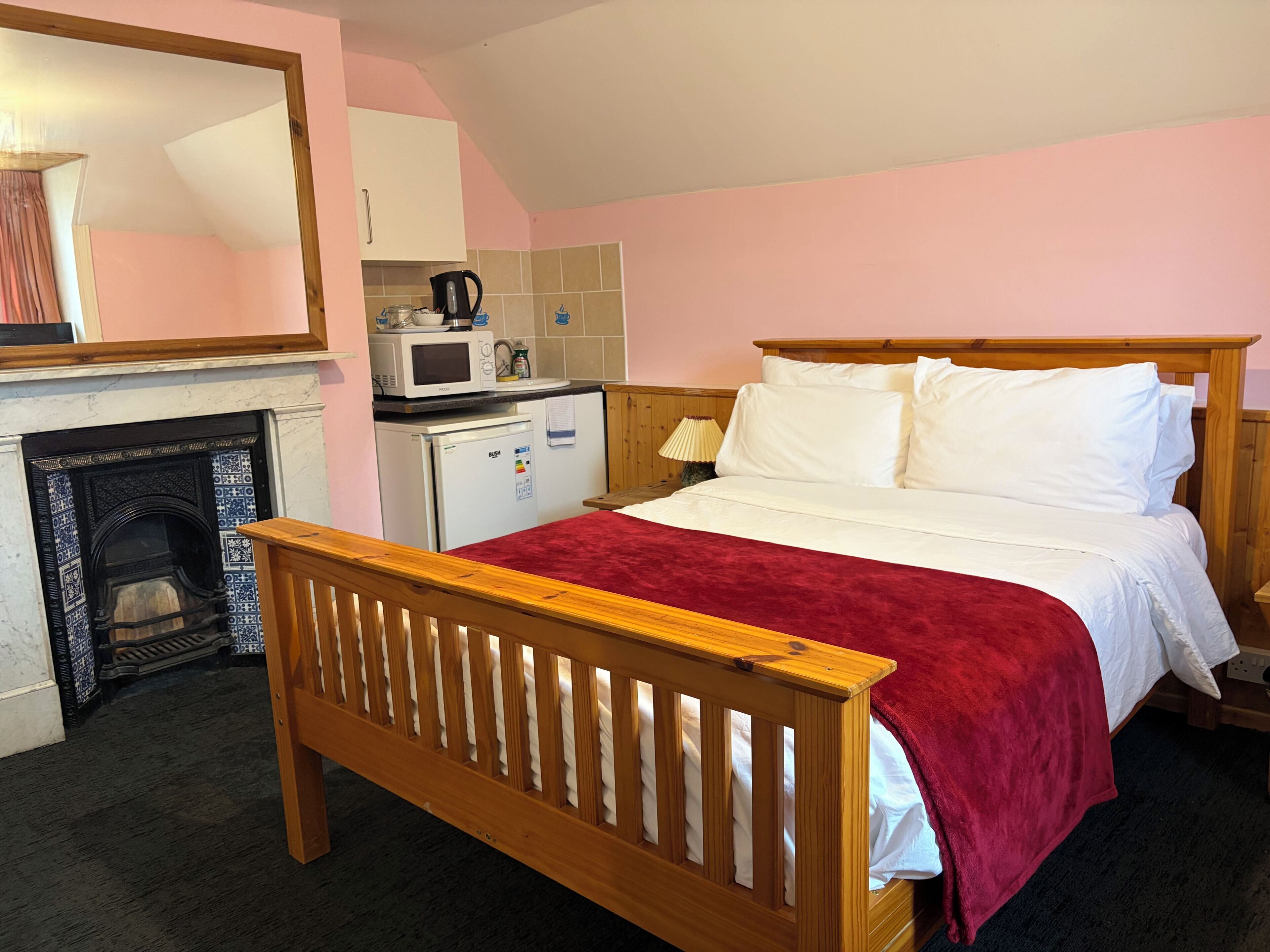 Ein Schlafzimmer mit einem hölzernen Bett, einer roten Decke, einem weißen Kopfteil, einer kleinen Küchenzeile mit Mikrowelle und Wasserkocher sowie einem Kamin.
