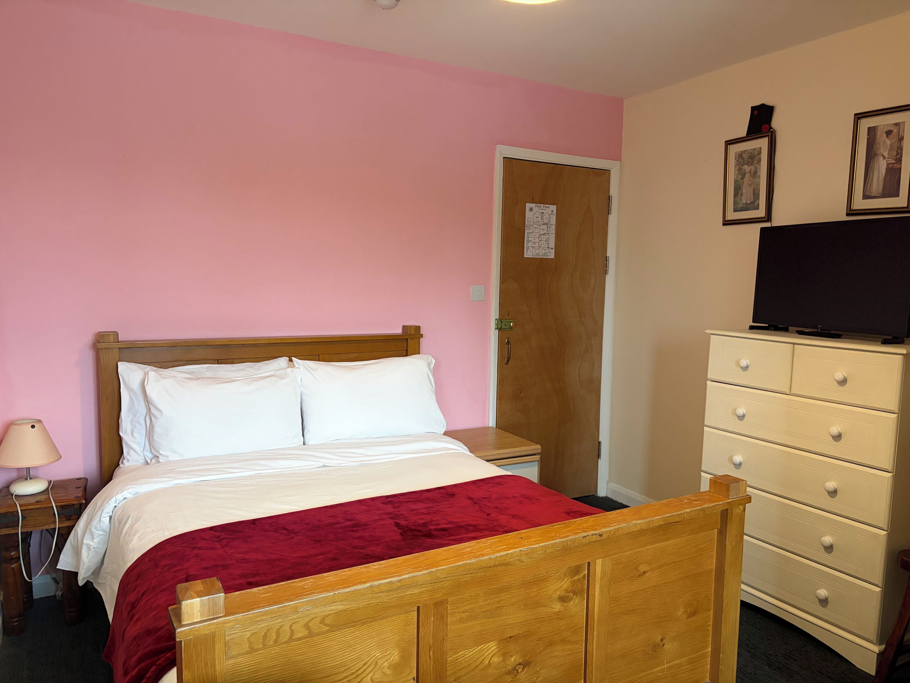 Une chambre à coucher avec un lit en bois, une couvre-lit rouge, une table de chevet avec une lampe, une commode avec des tiroirs et un téléviseur posé sur le dessus.
