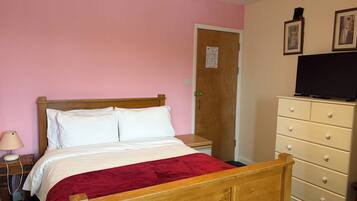 Une chambre à coucher avec un lit en bois, une couvre-lit rouge, une table de chevet avec une lampe, une commode avec des tiroirs et un téléviseur posé sur le dessus.