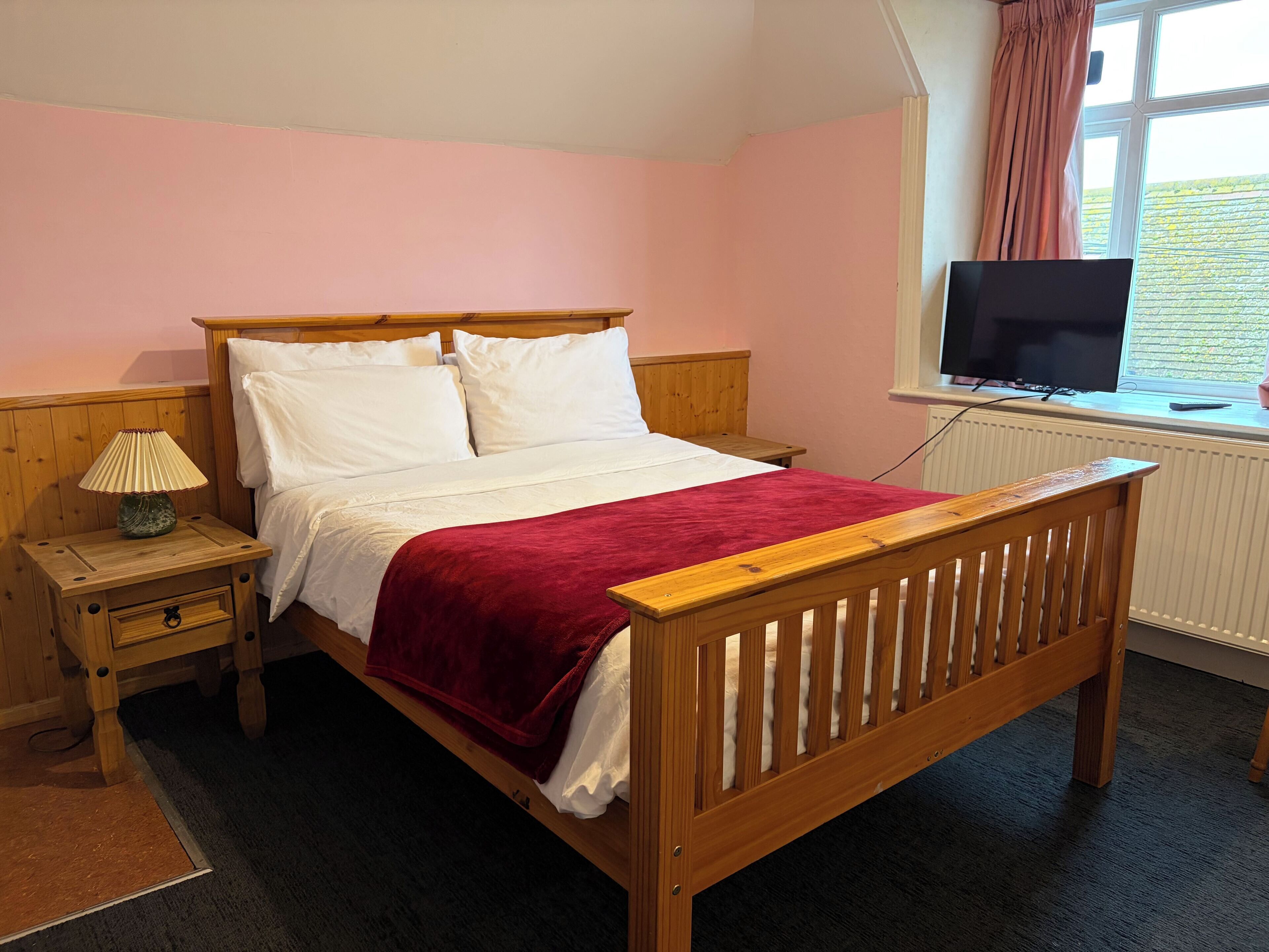 Une chambre avec un lit en bois, du linge de lit blanc, un plaid rouge, une table de chevet avec une lampe et un téléviseur posé sur le rebord d’une fenêtre.
