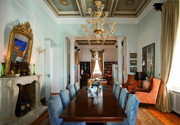 A formal dining room with a long table, blue upholstered chairs, a chandelier, and a fireplace. - Hotel Ploes (Syros)