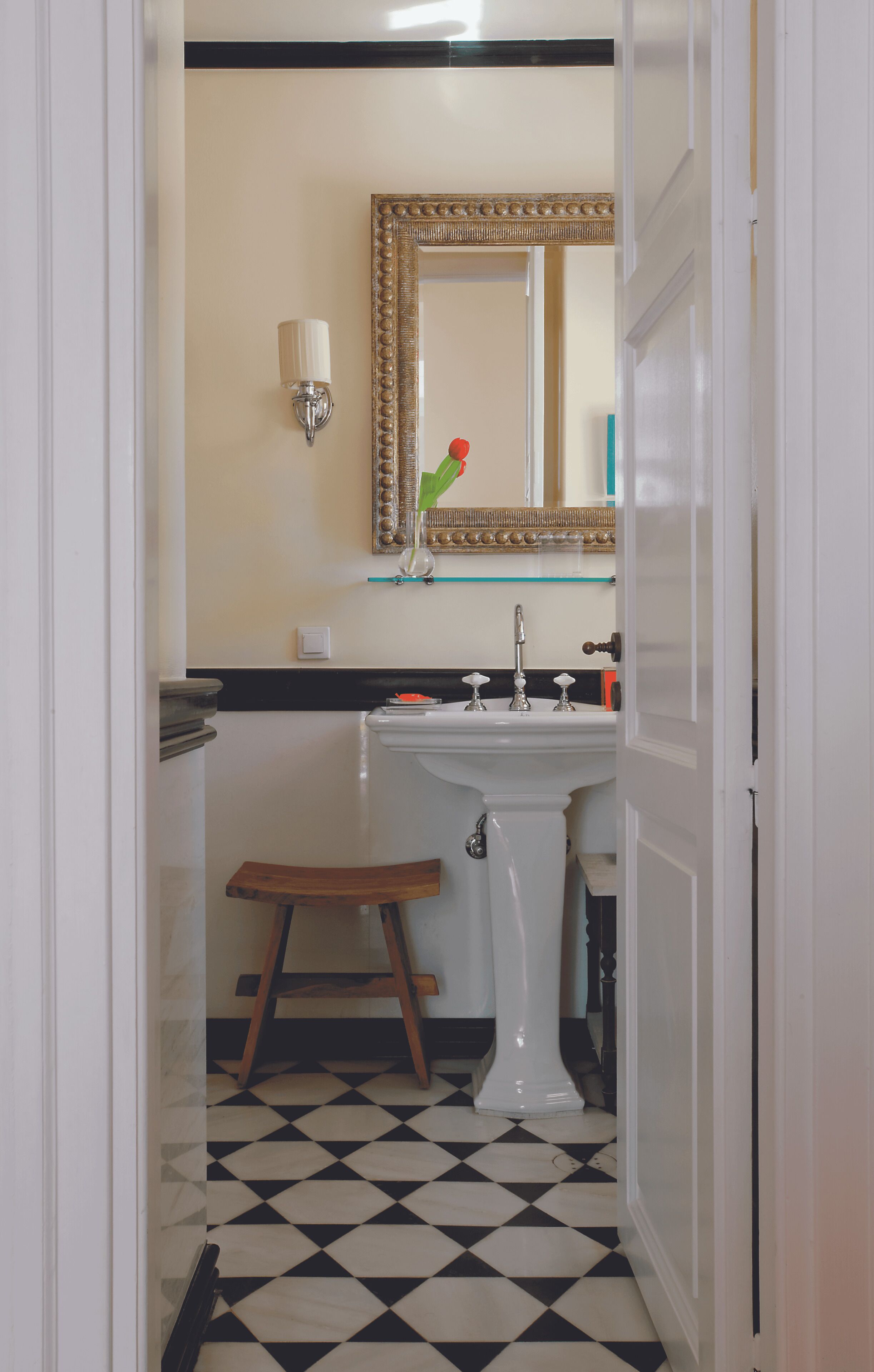 Um banheiro com pia branca, espelho com moldura decorativa, um banquinho pequeno e piso xadrez.