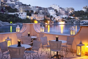 A terrace with wicker chairs and tables, lit by lanterns, overlooking a coastal town with buildings and a body of water.