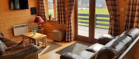 A cozy room with a sofa, a table with a lamp, a TV mounted on the wall, and a window with curtains.