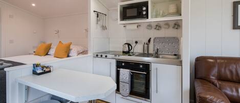 Een kleine keuken met een magnetron, fornuis en wastafel. Er is een eethoek met een tafel en twee krukken. Een bruine lederen stoel staat in de hoek.