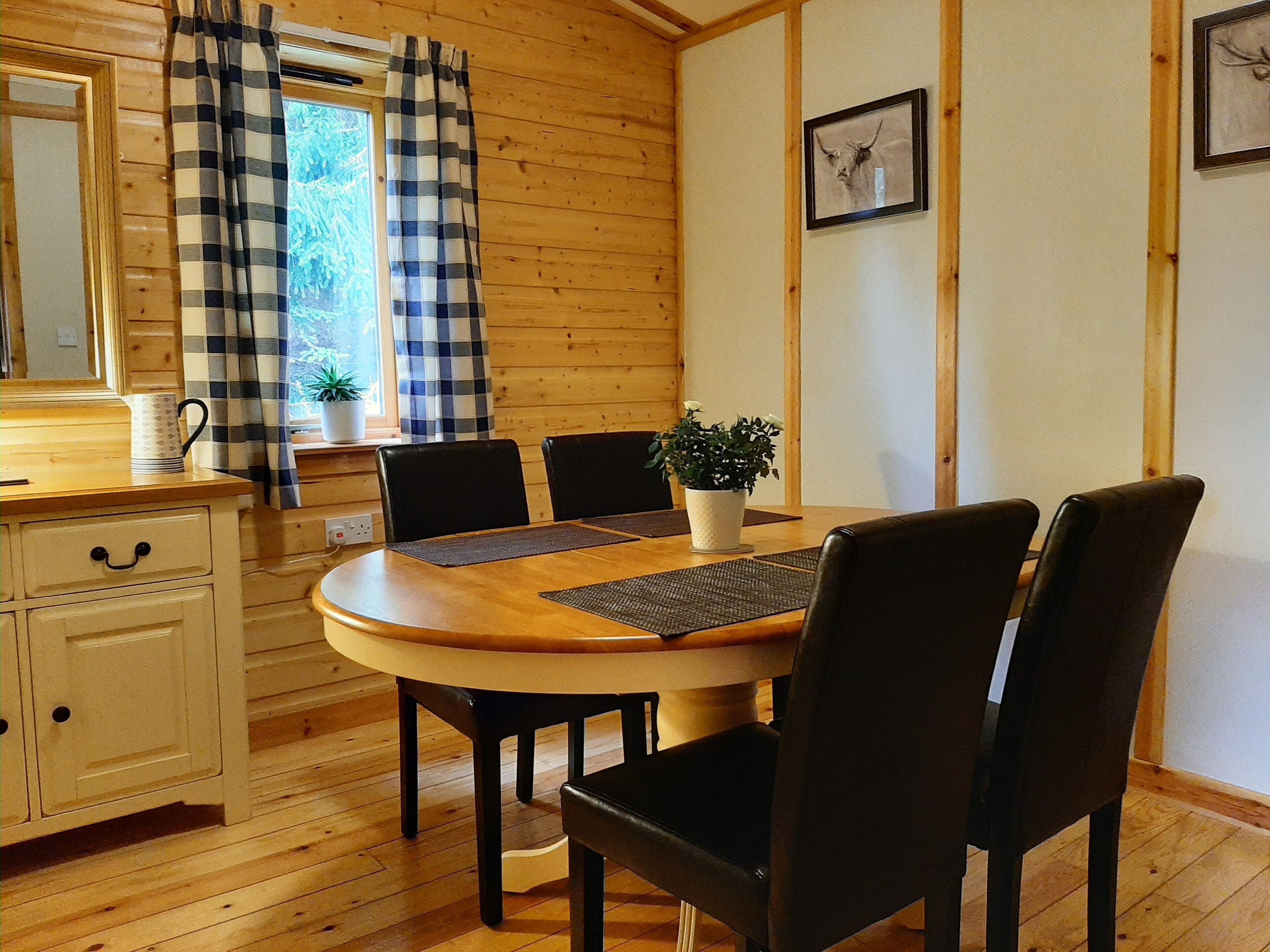 Un interior de madera con una mesa redonda, sillas negras, un armario blanco, una ventana con cortinas a cuadros y un cuadro en la pared.