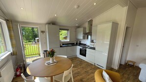 A compact kitchen with white cabinets, a dining table with chairs, a yellow sofa, and a view of a garden through a sliding door. - Tayview Lodges (Pitlochry)