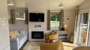 A cozy living room with a sofa, a yellow armchair, a fireplace, a TV mounted on the wall, and a window with curtains. - Tayview Lodges (Pitlochry)