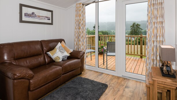 A brown leather sofa with patterned cushions, a grey rug, and a wooden deck with a view of greenery. - Tayview Lodges (Pitlochry)