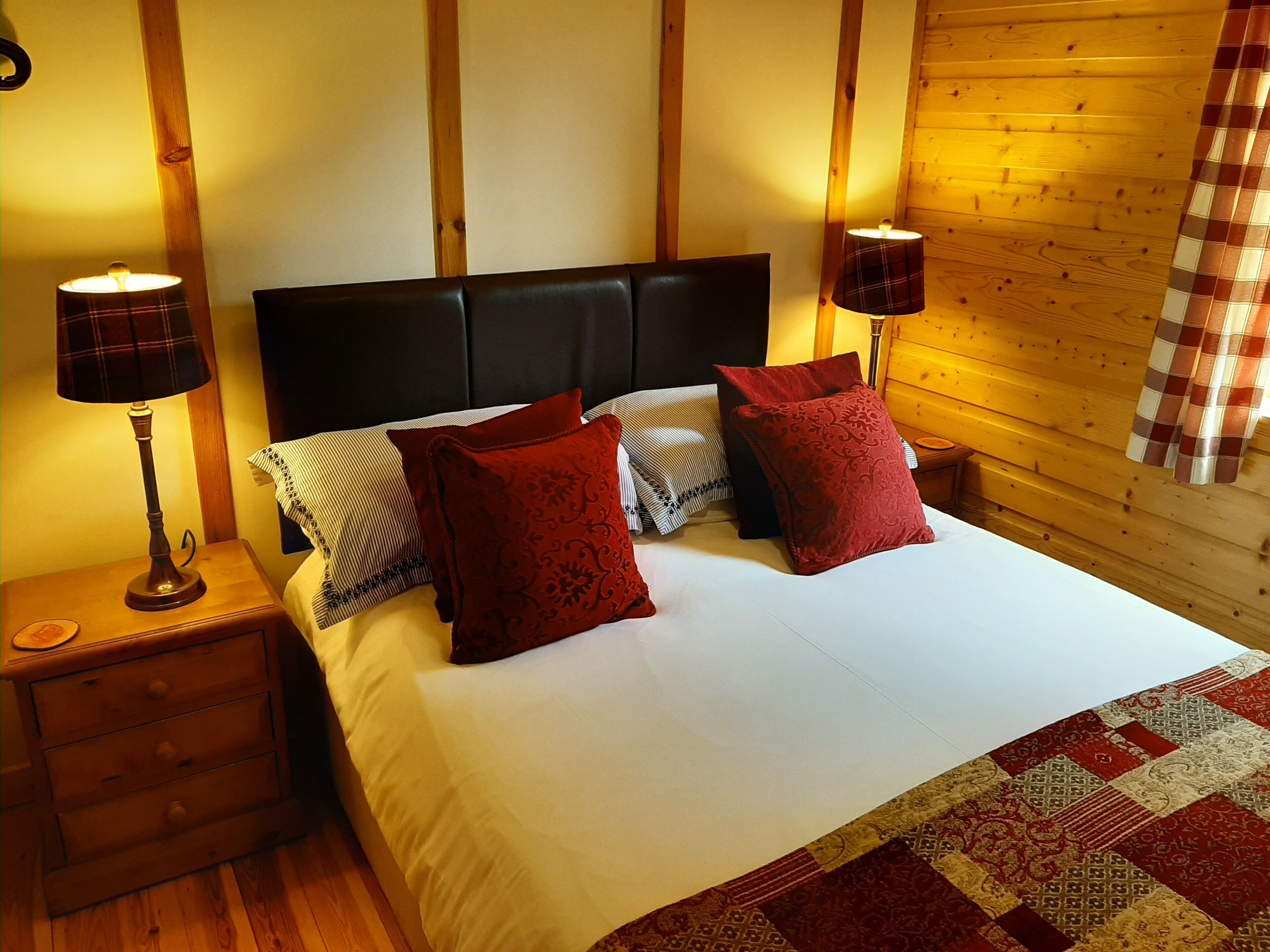 A neatly made bed with a white duvet and red pillows, flanked by two bedside tables with lamps, in a room with wooden walls and a plaid curtain.
