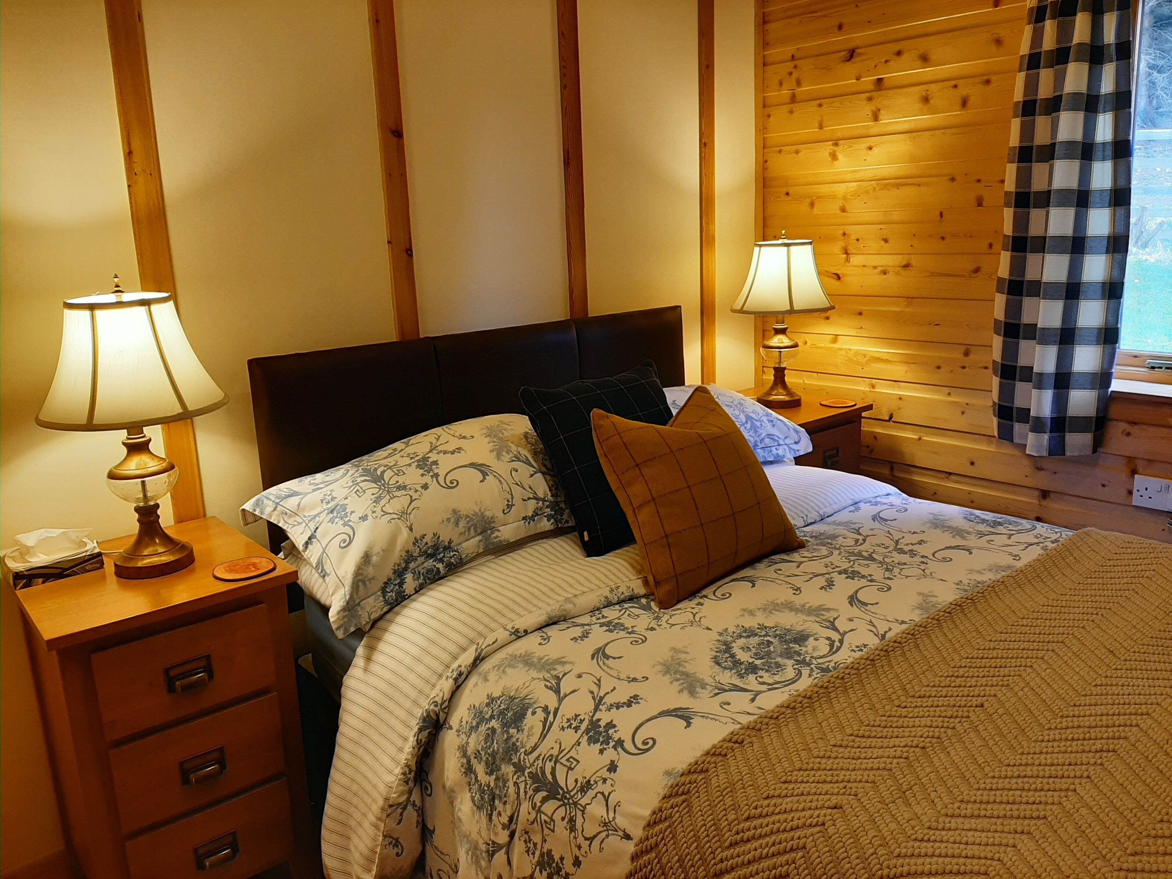 A neatly made bed with patterned bedding, two bedside lamps, a wooden nightstand with drawers, and a window with a checkered curtain.