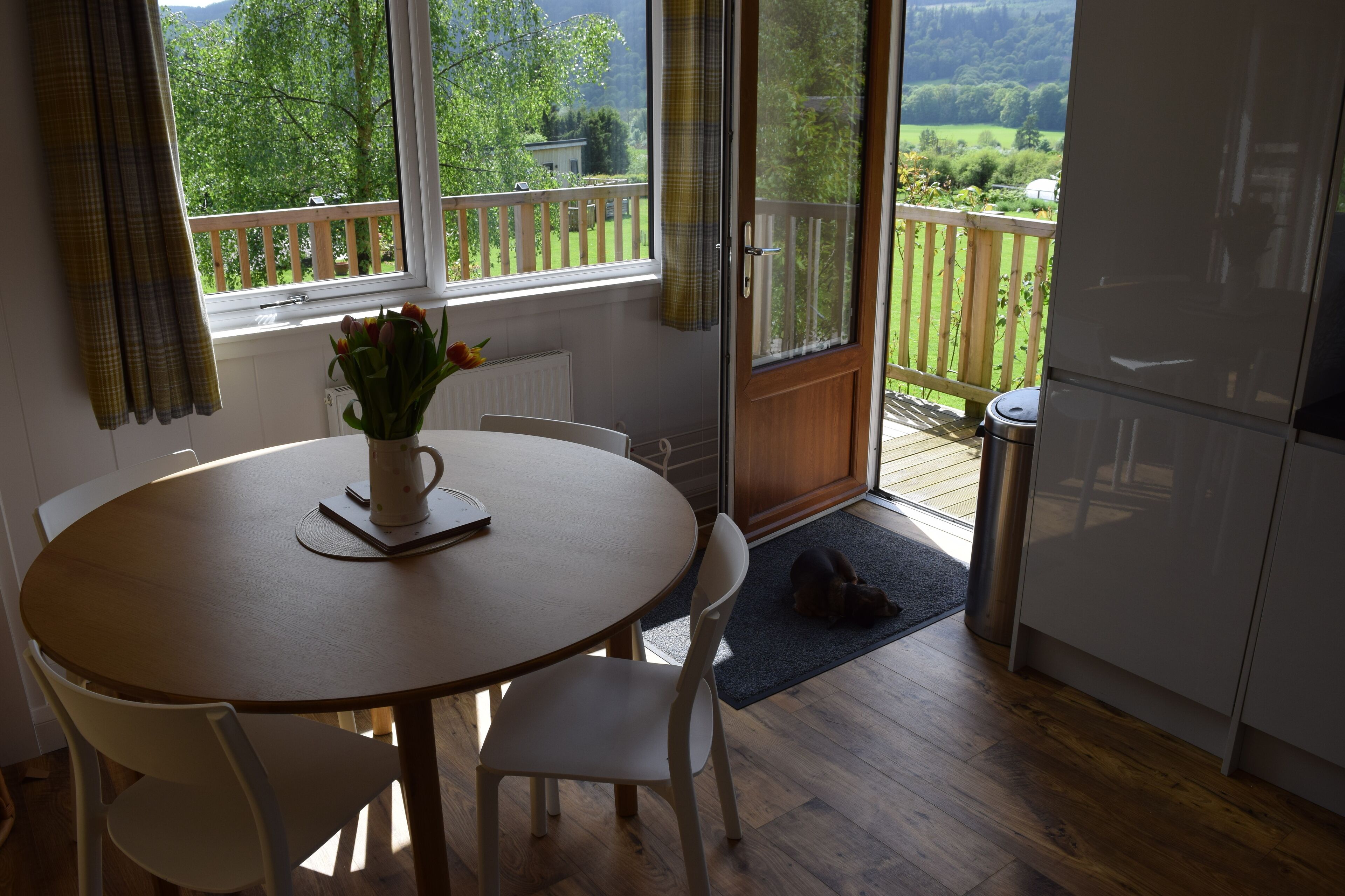 Un comedor con una mesa, sillas, un florero con flores, una ventana con vista a follaje y una puerta corrediza que da a una terraza exterior.