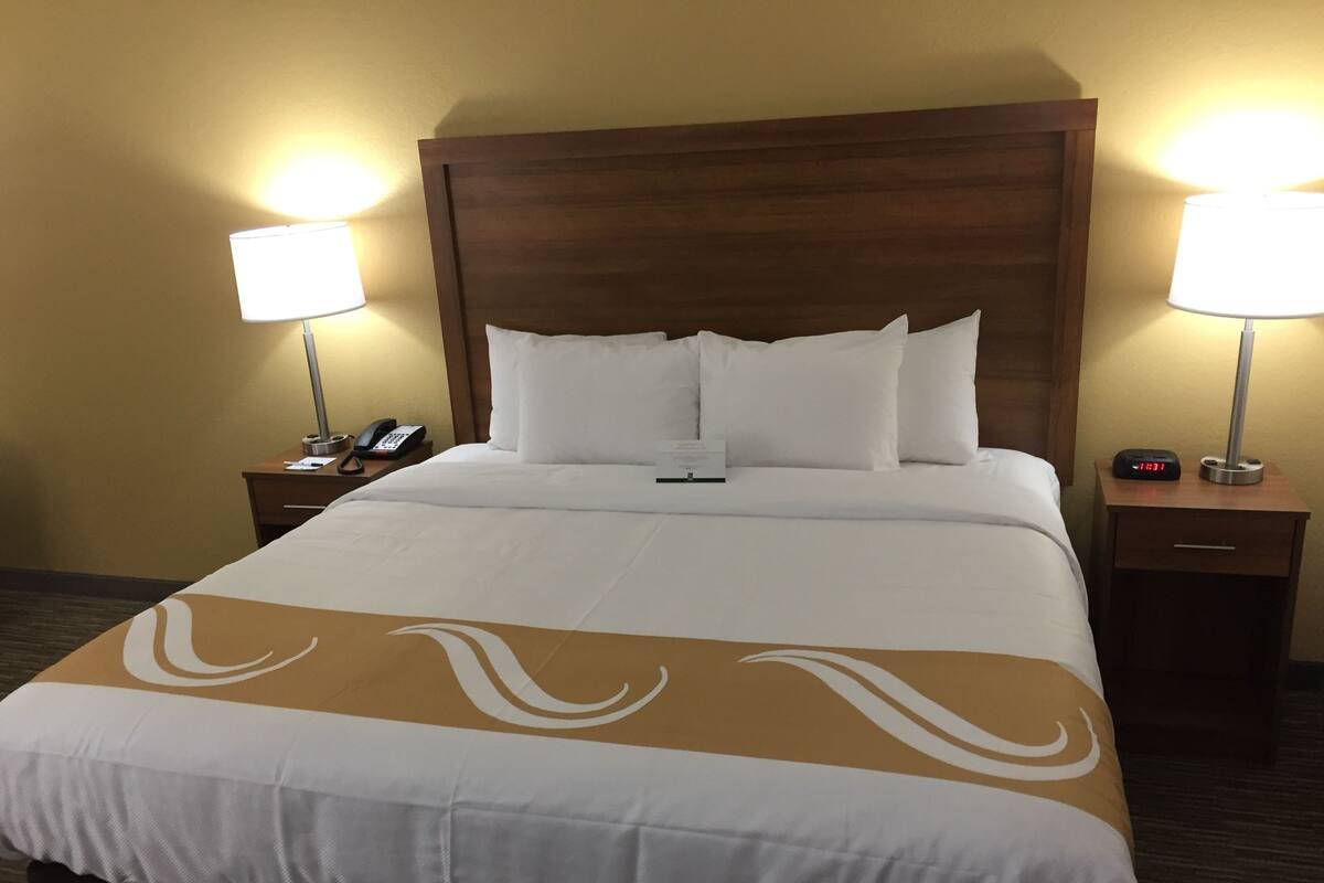 Habitación de hotel con una cama grande, dos mesitas de noche, una lámpara, un teléfono y un reloj digital.