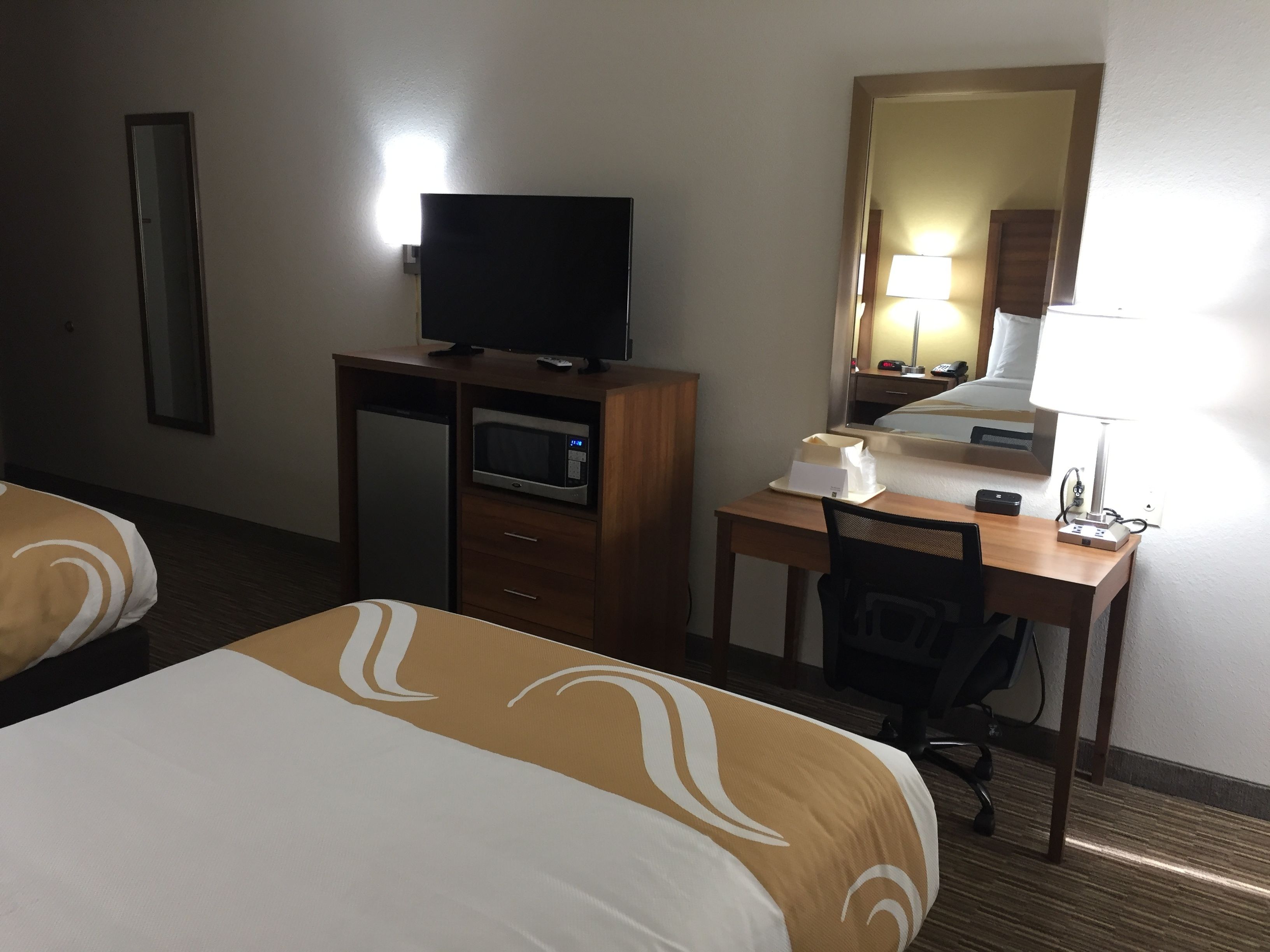 A hotel room with two beds, a desk with a computer, a TV, a microwave, and a bedside table with a lamp.