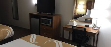A hotel room with two beds, a desk with a computer, a TV, a microwave, and a bedside table with a lamp.