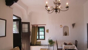 A cozy living room with a sofa, a dining table with chairs, a chandelier, a window with curtains, and a door. - Apartamentos Rurales Las Caleras (Almagro)
