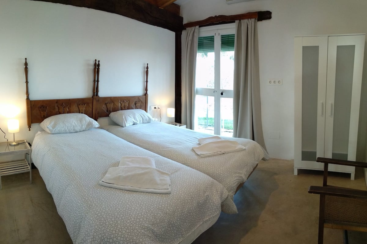 A double bed with a wooden headboard, two bedside tables with lamps, a wardrobe, and a view of the outdoors through a window with curtains.