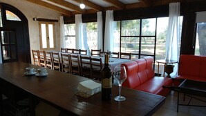 A dining area with a wooden table, chairs, a bottle of wine, and a glass. There is a red sofa and a small round table with a lamp. Large windows with curtains are visible. - Apartamentos Rurales Las Caleras (Almagro)