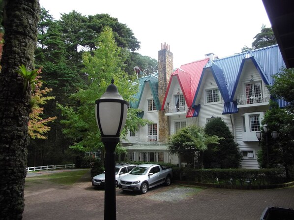 A building with a stone chimney, multiple windows, and a paved driveway with parked cars. - Canada Lodge (Campos do Jordão)