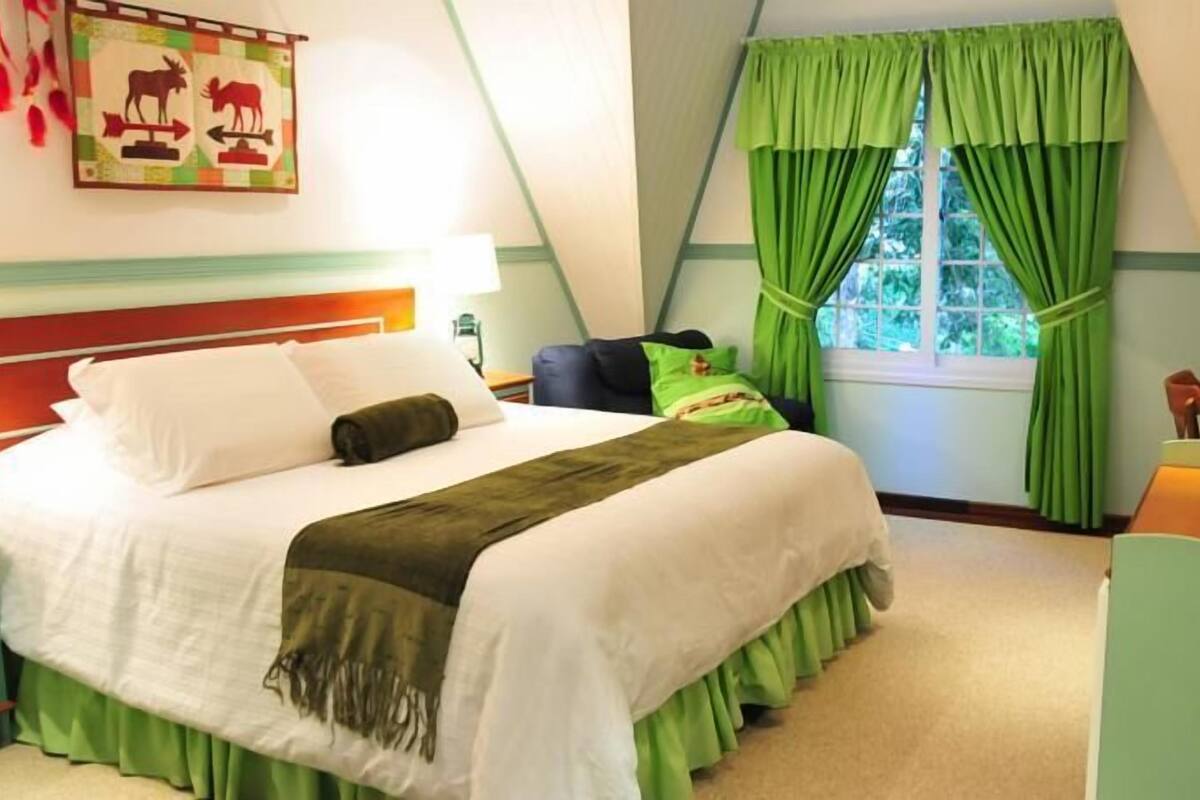 Hotellihuone, jossa on sänky, vihreät verhot, ikkuna ja pieni pöytä.