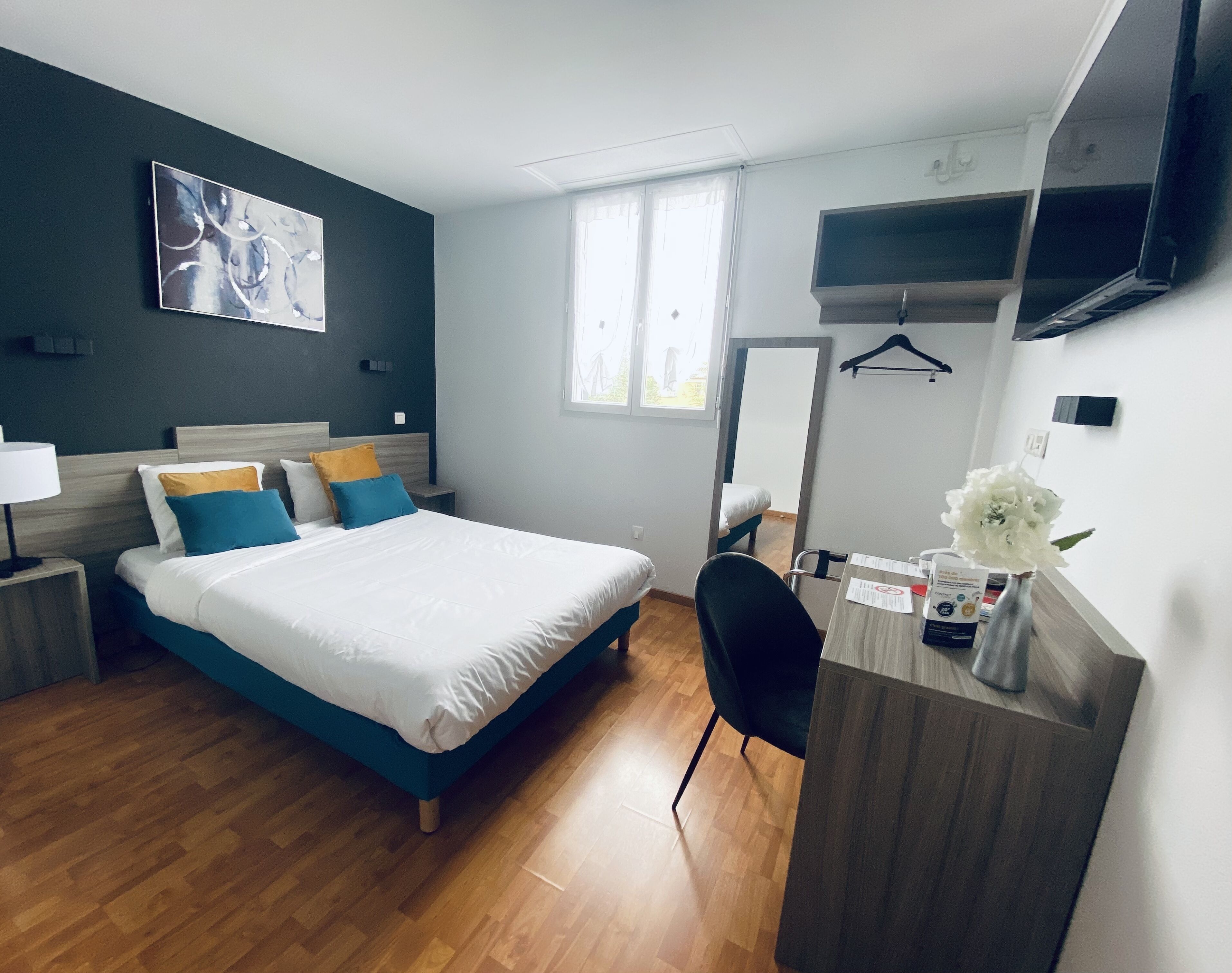 A hotel room with a bed, bedside table, chair, desk, mirror, and a TV on the wall.