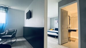 Une chambre d’hôtel moderne, équipée d’un lit, d’un coin salon avec un fauteuil, d’une télévision et d’une salle de bain visible à travers une porte ouverte.