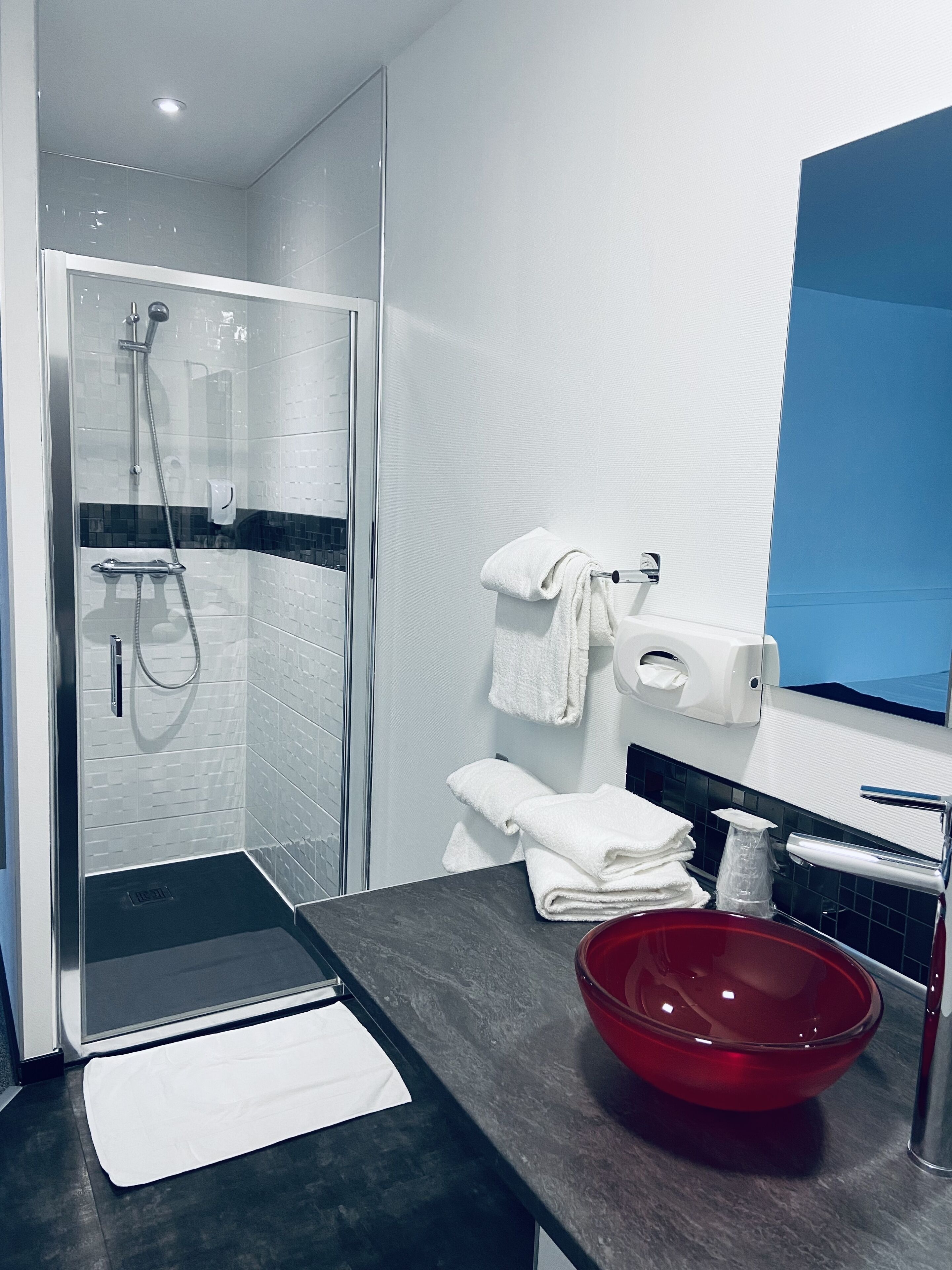 A modern bathroom with a glass-enclosed shower, a white towel rack, a red bowl sink, and a blue accent wall.