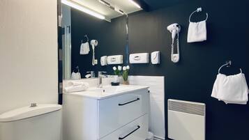 Een moderne badkamer met een wit toilet, een witte wastafel en een spiegel boven de wastafel. Handdoeken hangen aan haken en een haardroger zit aan de muur.