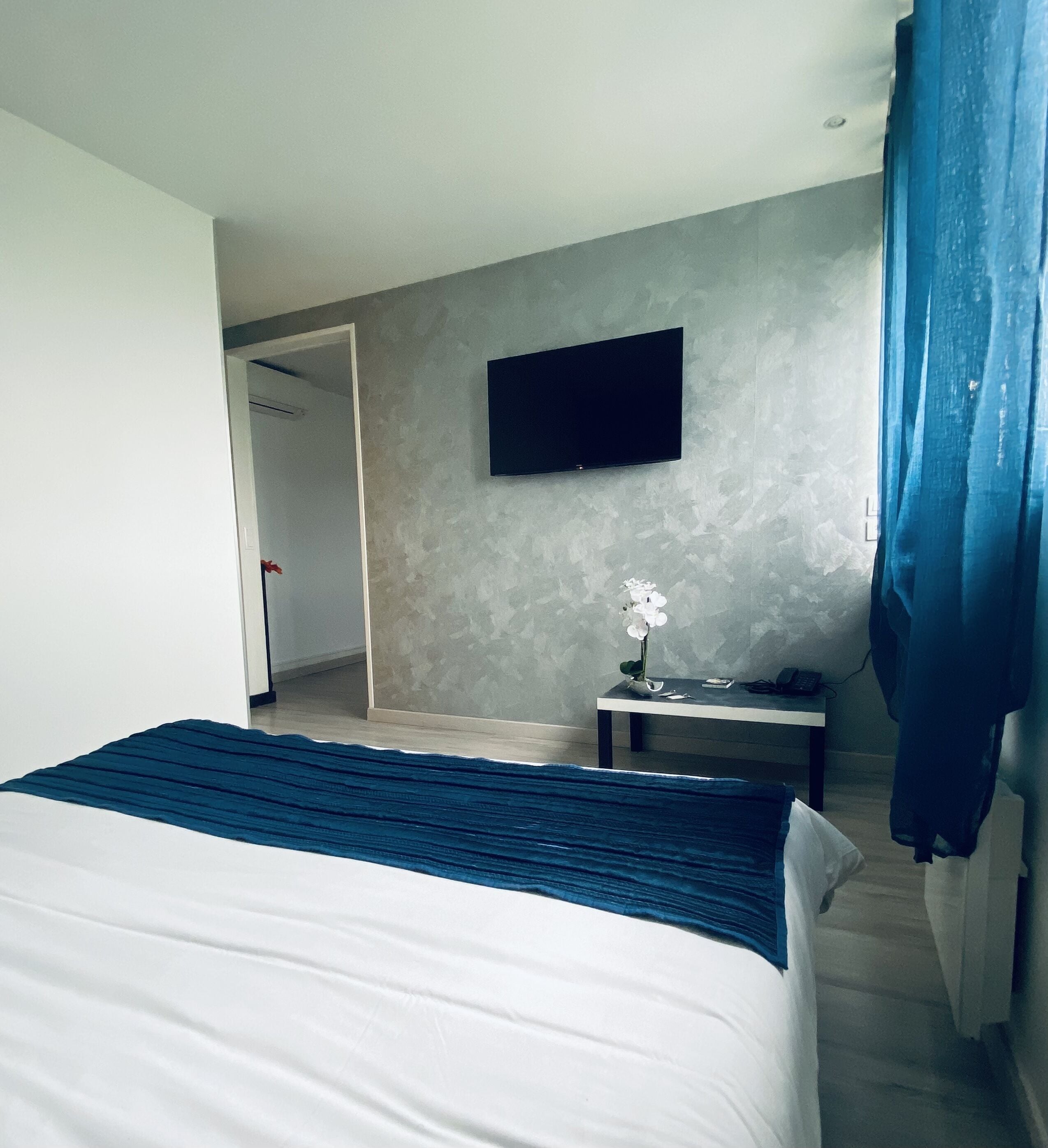 Quarto de hotel com cama, televisão de tela plana na parede, mesa pequena com um vaso de flores e cortina azul.