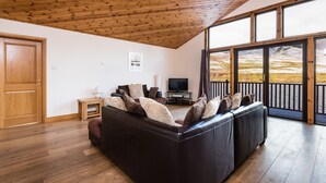 A living room with a dark leather sofa, wooden flooring, a television, and a large window with a view of a snowy landscape. - Scottish Highland Lodge (Pitlochry)