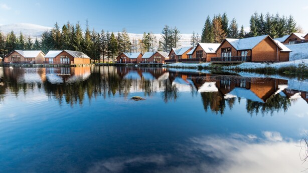 A row of log cabins by a calm lake with reflections, surrounded by snow-covered ground and trees. - Scottish Highland Lodge (Pitlochry)