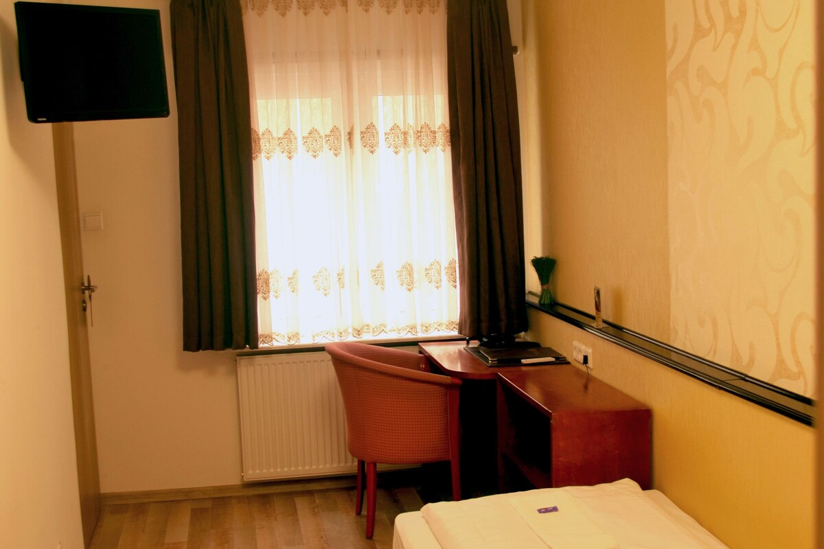 Quarto de hotel com cama, escrivaninha, cadeira, televisão fixada na parede e janela com cortinas.