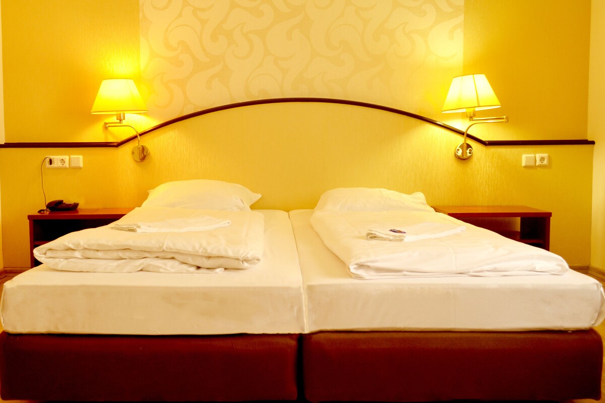Quarto de hotel com duas camas, cada uma com lençóis e almofadas brancas, cabeceira de madeira e duas luminárias de cabeceira.